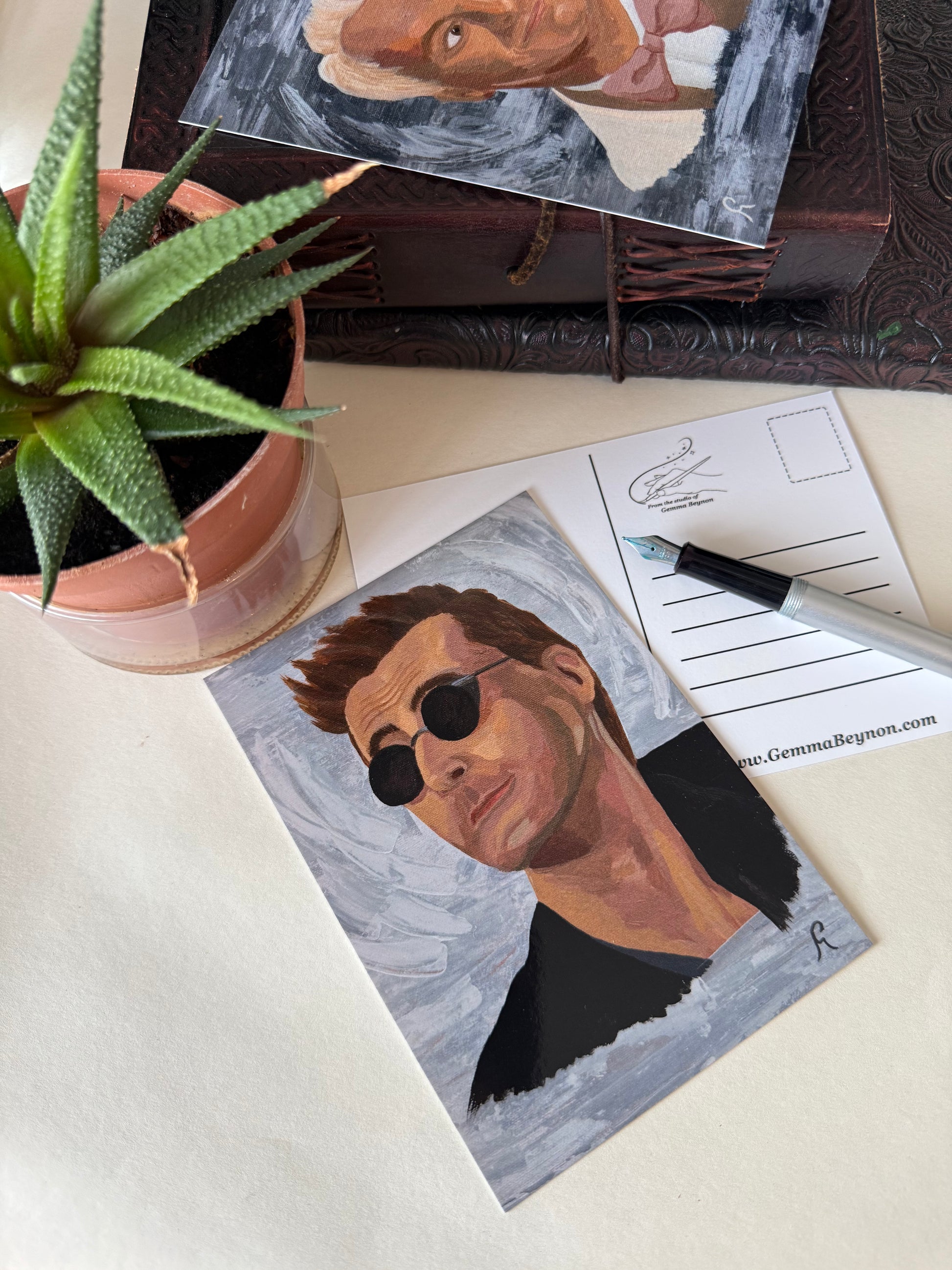 Artistic postcard portrait of a Crowley  wearing sunglasses on a table with a plant and stationery items.